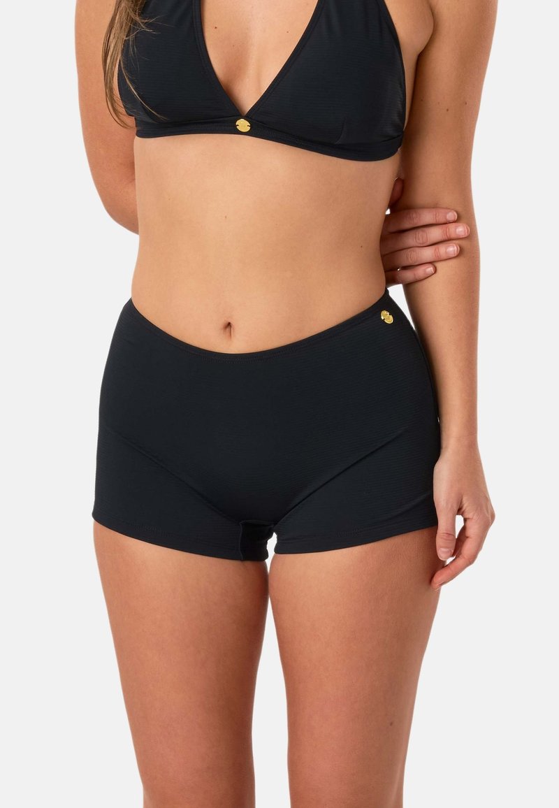 Femme portant un short de bain noir taille haute et un haut de bikini assorti, avec un bras plié et la main reposant sur le bras opposé.