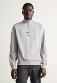 Světlá šedá mikina na zip s vysokým límcem, žebrovanými manžetami a spodním lemem. Na přední straně je modrý detail s logem. Hladká textura látky.