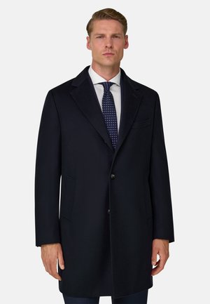 Homme portant un pardessus formel sombre, une chemise blanche et une cravate sombre à motifs, debout devant un fond clair uni.