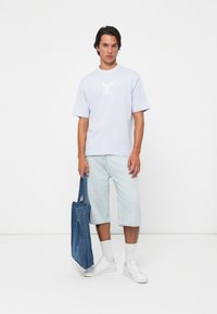 Mann som har på seg en lys blå t-skjorte og denimshorts til kneet, holder en blå denim tote bag, står mot en hvit bakgrunn.