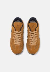 Tenis de camurça castanhos com atacadores bege, laterais texturizadas e um logo preto. Bico arredondado e sola branca com detalhes pretos.