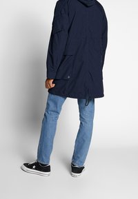 Mörkblå parka jacka med huva, framfickor och dragsko i nederkant, kombinerad med ljusblå jeans och svarta sneakers med stjärndetalj.