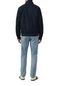 Marine bomberjack met een hoge kraag, geribbelde manchetten en zoom, gecombineerd met lichtblauwe rechtpijpen jeans en witte sneakers met bruine accenten.