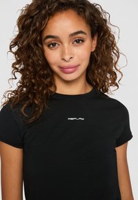 Jonge vrouw met krullend bruin haar die een zwart Replay T-shirt draagt, vriendelijk glimlachend tegen een eenvoudige lichte achtergrond.
