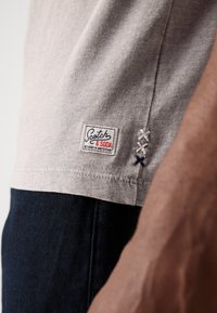 Light gray cotton t-shirt featuring a logo patch and decorative stitching in white and navy, with a classic fit and a curved hem.