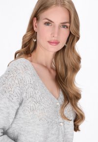 Cardigan gri tricotat cu decolteu în V, având un model din dantelă și butoni din perlă. Modelul are părul lung și ondulat și poartă cercei în formă de lacrimă.