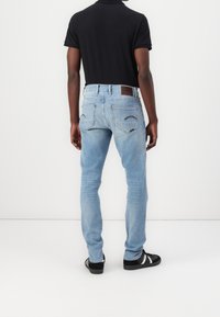 Vaqueros de color azul claro con un corte ajustado, que cuentan con dos bolsillos traseros y un parche de cuero marrón. Combinados con una camiseta polo negra y zapatos negros.