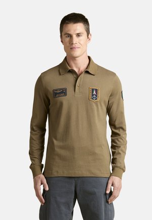 Uomo con capelli corti e scuri che indossa una polo marrone a maniche lunghe con due toppe ricamate e pantaloni grigio scuro, in piedi su uno sfondo bianco.
