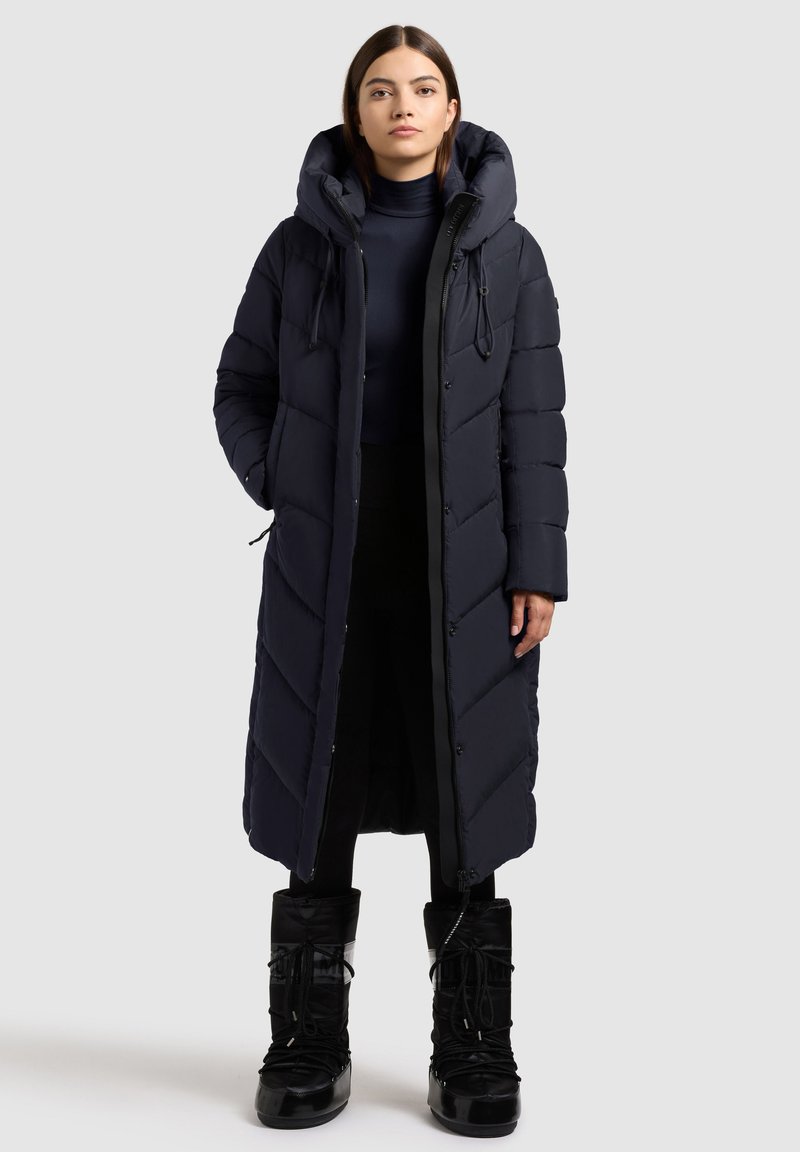 Lang, pufferjakke i mørk navy med hætte, med kviltet design, lynlås foran og sidelommer. Kombineret med sorte snestøvler.