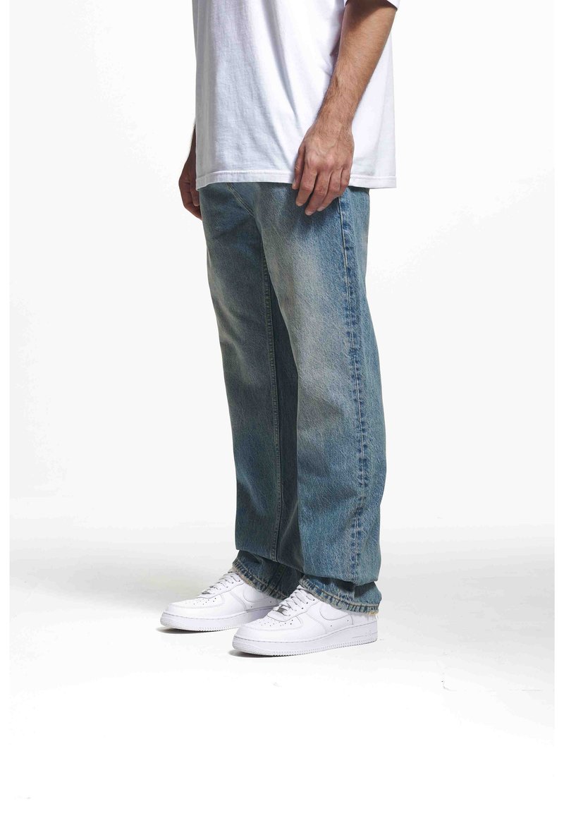 Jean en denim bleu clair avec une coupe décontractée, associé à des baskets blanches. Le jean présente un léger délavage et un design classique à cinq poches.