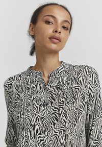 Blusa a stampa zebra in bianco e nero con colletto alla mandarina, dettagli plissettati e tessuto leggero. Maniche lunghe e vestibilità comoda.