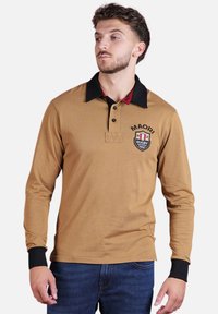 Polo à manches longues marron avec un col noir, des accents rouges et un emblème de rugby portant "MAORI". Présente des poignets et des coutures contrastants en noir.
