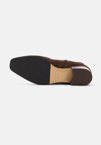 Zapato de ante marrón con suela de goma plana, que presenta un ligero tacón y un diseño limpio y minimalista. Detalle de costura notable a lo largo de la parte superior.