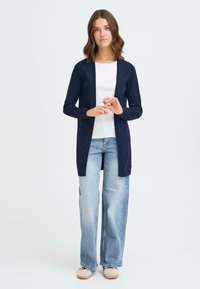 Un cardigan marine en tricot léger, à l'avant ouvert, associé à un haut blanc à côtes et un jean large bleu clair. Chaussures de teintes neutres.