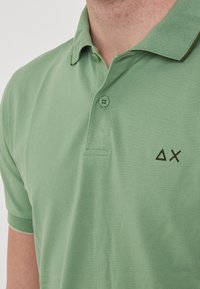 Polo shirt verde realizzato in tessuto texturizzato, con una patta a due bottoni e un piccolo logo marrone sul petto. Maniche corte.