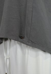 Top grigio in cotone con orlo arrotondato, caratterizzato da un piccolo logo. Indossato sopra un indumento bianco con dettaglio vita con cordino.
