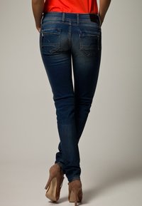 Kvinna i mörkblå slim fit jeans och beige högklackade skor, stående med benen korsade, synlig bakifrån.