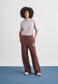 Ανοιχτό μοβ κοντομάνικο πουκάμισο με λογότυπο "NIKE SHOX", συνδυασμένο με φαρδύ καφέ παντελόνι φόρμας με λευκό λογότυπο Nike και κορδόνι μέσης. Χαλαρά αθλητικά παπούτσια.