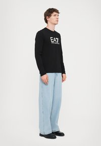 Fekete, hosszú ujjú póló "EA7 EMPORIO ARMANI" logóval, világoskék, bő szárú farmernadrággal és fekete cipőkkel párosítva. Egyszerű és laza stílus.