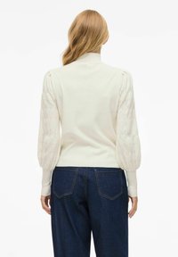 Mujer rubia de espaldas, vestida con un suéter de punto color crema con mangas abullonadas y estampadas, y jeans oscuros, frente a un fondo blanco.