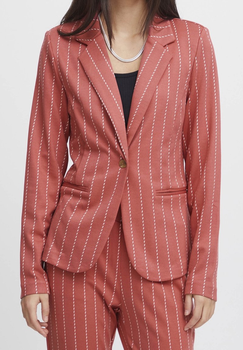 Femme portant un costume rayé couleur rouille avec un blazer à un seul bouton et un pantalon assorti, sur un top noir et un collier argenté.