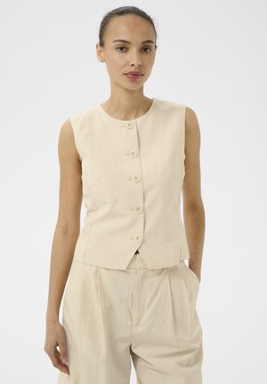 Kobieta z włosami zaczesanymi do tyłu, ubrana w bezrękawnik w kolorze beżowym zapinany na guziki oraz dopasowane, luźne spodnie z wysokim stanem na jasnym tle.