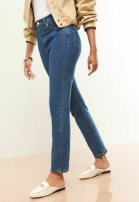 Blauwe denim jeans met een rechte snit, hoge taille en subtiele stikdetails. Gecombineerd met witte instap schoenen op een lichte achtergrond.