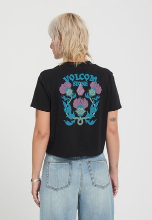 Personne aux cheveux blonds portant un t-shirt court noir Volcom Stone avec un motif floral bleu et rose dans le dos et un jean taille haute bleu clair.