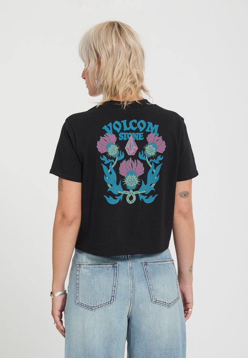 Personne aux cheveux blonds portant un t-shirt court noir Volcom Stone avec un motif floral bleu et rose dans le dos et un jean taille haute bleu clair.