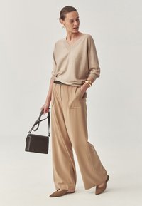 Beige svetr s výstřihem do V z vlněné směsi, který je spárován s béžovými kalhotami s širokými nohavicemi. Doplňky zahrnují černou kabelku a zlatý náramek. Měkká textura je zřejmá.