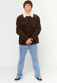 Brun kordfløjljakke med en hvid pelskrave, to brystlommer og et firkantet logo patch. Båret med lyseblå jeans og sorte sneakers.