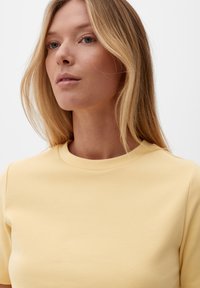 Blondynka z prostymi włosami, ubrana w jasną żółtą bluzkę z okrągłym dekoltem, patrzy nieco na bok na jednolitym tle.