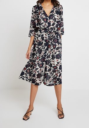 Kobieta ubrana w sukienkę kopertową o długości do kolan, w kwiatowy wzór, z rękawami 3/4 i paskiem wiązanym w talii, w parze z czarnymi sandałami na obcasie z paskami.