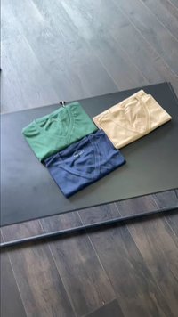 Tre t-shirt a scollo a V: una verde, una blu navy e una beige, disposte ordinatamente su una superficie nera piatta. Materiale in cotone, etichette visibili.