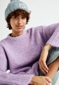 Lila vuna džemper s rebrastim manžetama, opuštenog kroja i meke teksture. Uparen s sivom pletenom kapom i ispucanim trapericama.