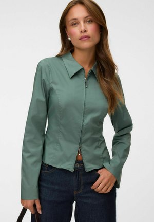 Femme aux longs cheveux bruns portant une veste verte ajustée zippée, un jean bleu foncé, des boucles d'oreilles en perles et tenant un sac à bandoulière noir.