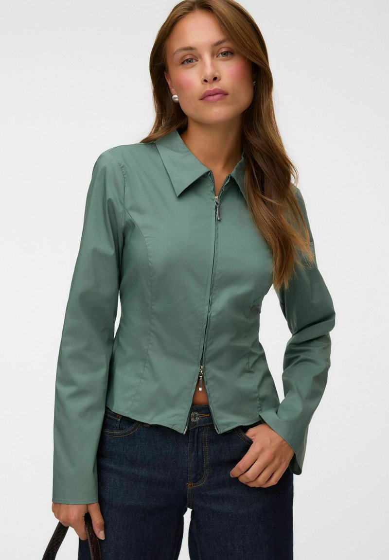 Femme aux longs cheveux bruns portant une veste verte ajustée zippée, un jean bleu foncé, des boucles d'oreilles en perles et tenant un sac à bandoulière noir.