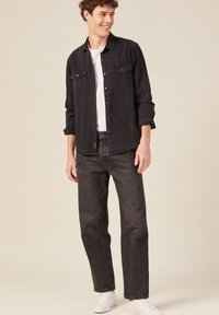Camisa de denim negra con dos bolsillos en el pecho, botonadura frontal y mangas largas sobre una camiseta blanca. Combinada con jeans oscuros y zapatillas deportivas blancas.