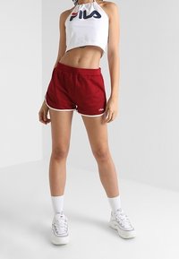 Mujer vestida con un top corto halter blanco de FILA, pantalones cortos deportivos rojos de FILA con ribete blanco, calcetines blancos y zapatillas blancas, de pie contra una pared sencilla.