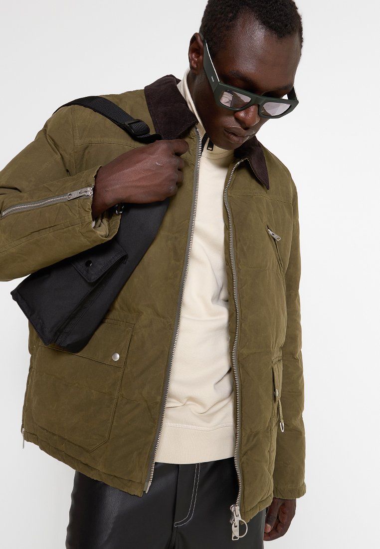 Oliwkowa, pikowana kurtka z czarnym kołnierzem, srebrnymi zamkami i licznymi kieszeniami. Nosi ją na jasnobeżowej bluzce z czarnymi spodniami.