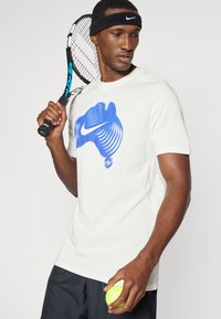 Λευκό βαμβακερό T-shirt με μπλε λογότυπο Nike, συνδυασμένο με μαύρη βερμούδα, κρατώντας ένα ρακέτα τένις και μια νεόν πράσινη μπάλα.