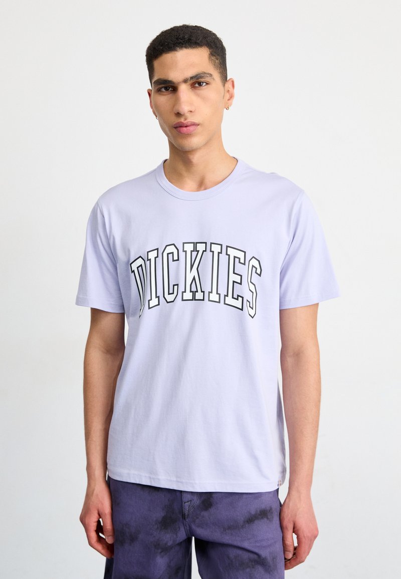Levandulové bavlněné tričko s zakřiveným černým potiskem loga "DICKIES" na hrudi. Krátké rukávy a kulatý výstřih.