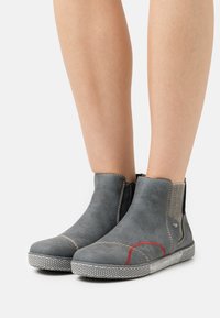 Bottines en suède gris avec panneaux latéraux élastiques, surpiqûres rouges contrastantes et semelle en caoutchouc blanc texturé.