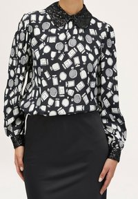 Blusa nera con stampa di bottiglia di profumo bianca, dotata di colletto nero con paillettes e maniche lunghe testurizzate. Abbinata a una gonna a tubino nera.