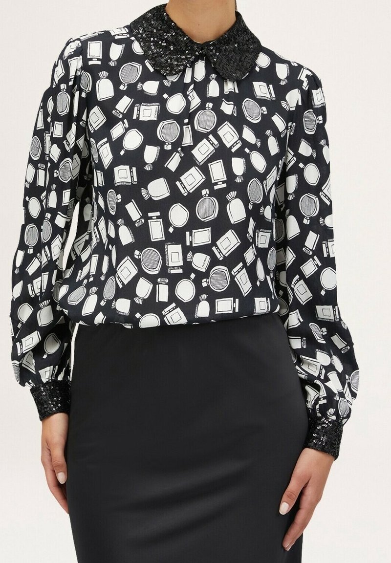 Blusa nera con stampa di bottiglia di profumo bianca, dotata di colletto nero con paillettes e maniche lunghe testurizzate. Abbinata a una gonna a tubino nera.