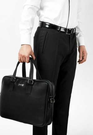 Muž v černých kalhotách a bílé košili drží černou koženou aktovku s kovovým logem na přední straně.