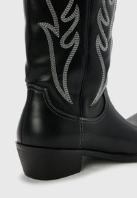 Bota de vaquero de cuero negro con bordados plateados en un patrón de llamas. Presenta un tacón bajo apilado y detalles de costura blanca en contraste.