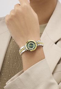 Καρπός που φοράει ρολόι Guess με χρυσή και ασημένια μπάντα, πράσινο καντράν και κρυστάλλινους δείκτες, πάνω από μπεζ σακάκι και υφασμένο πουκάμισο.