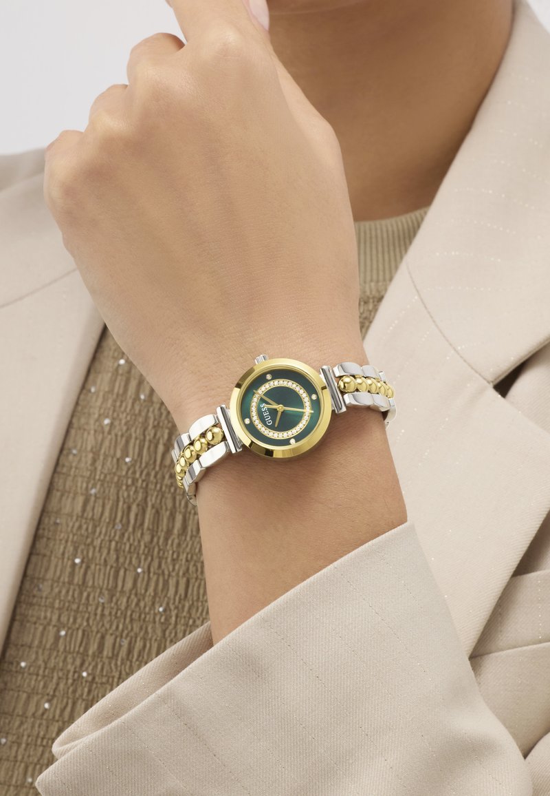 Καρπός που φοράει ρολόι Guess με χρυσή και ασημένια μπάντα, πράσινο καντράν και κρυστάλλινους δείκτες, πάνω από μπεζ σακάκι και υφασμένο πουκάμισο.
