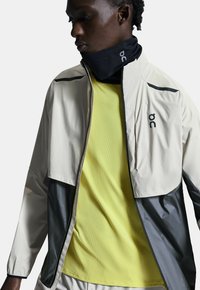 Veste légère beige et grise, avec un col haut, des accents noirs et un logo. Portée sur une chemise jaune texturée. Design doux et fonctionnel.